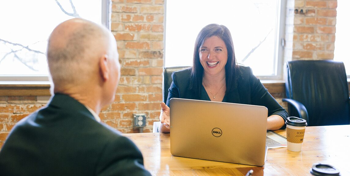 A woman in a dark blazer smiles while talking to a man across a wooden table with a Dell laptop and two paper coffee cups in a brick office.
