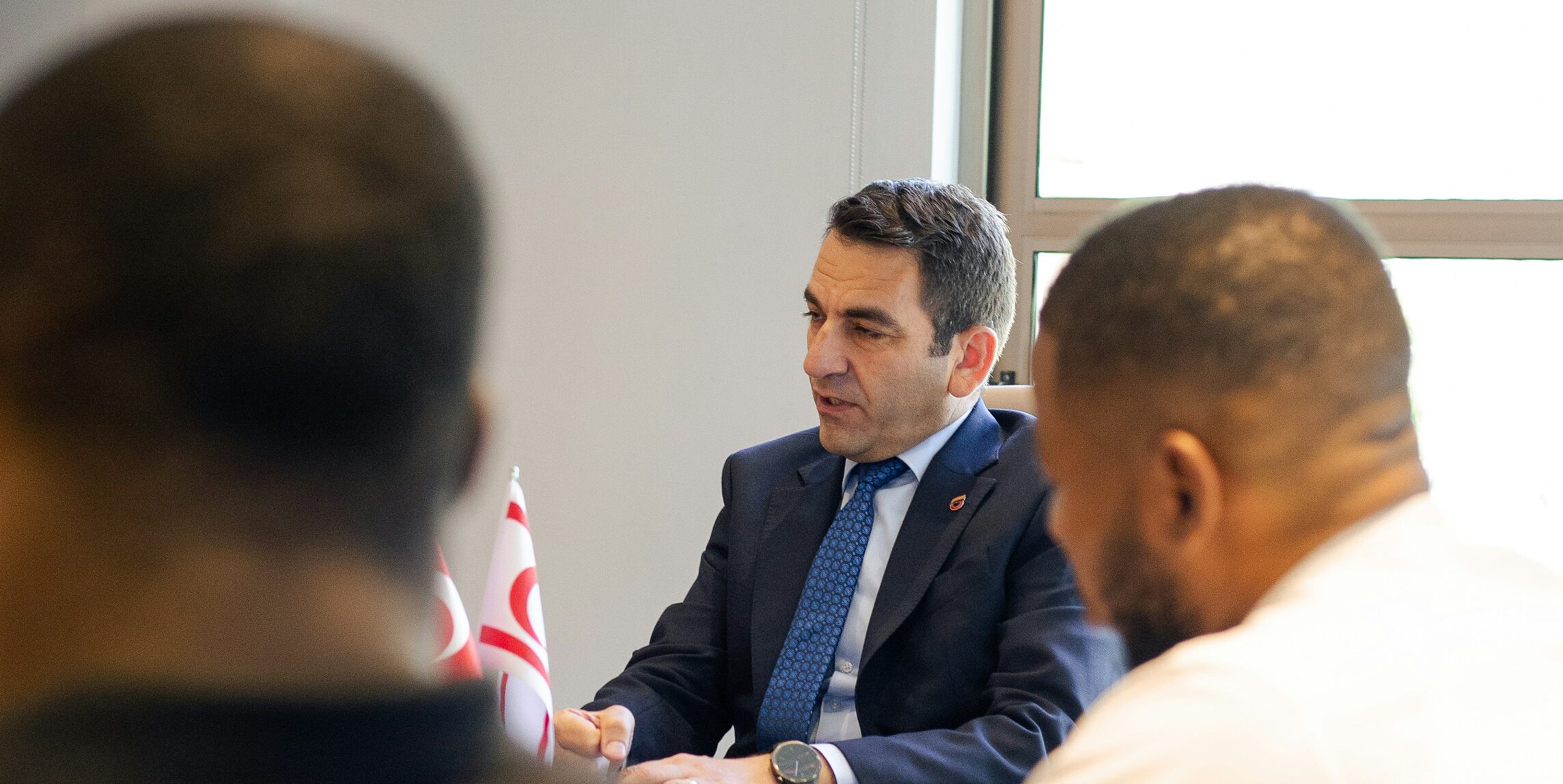 A man in a suit and tie sits at a table during a meeting, with two colleagues in the foreground and small flags visible on the wooden desk surface.