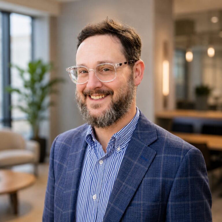 Man with a beard and glasses, smiling, standing in a lounge area with soft seating in the background