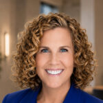 Woman with curly hair in a blue blazer, smiling, standing in an open office area near a conference table