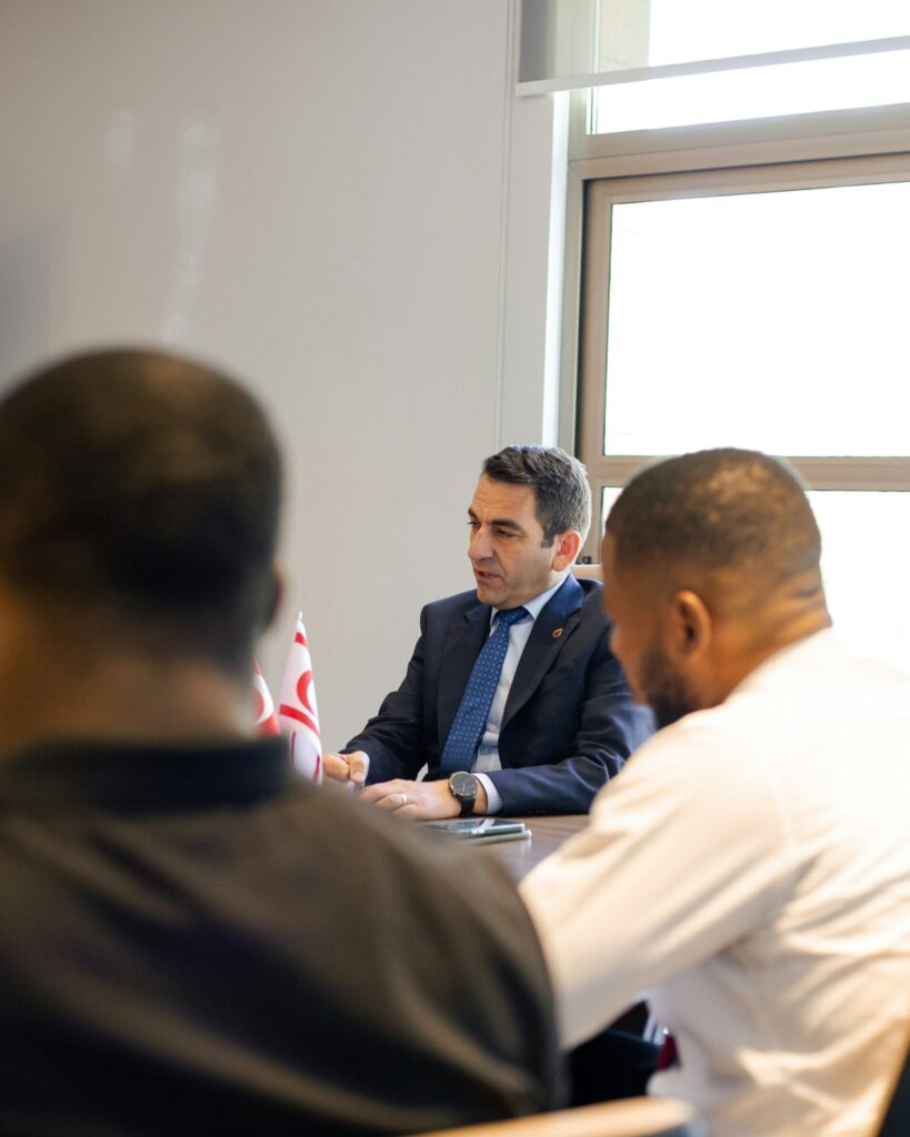 A man in a suit and tie sits at a table during a meeting, with two colleagues in the foreground and small flags visible on the wooden desk surface.