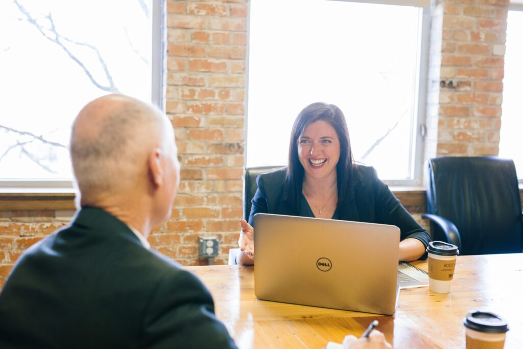 A woman in a dark blazer smiles while talking to a man across a wooden table with a Dell laptop and two paper coffee cups in a brick office.