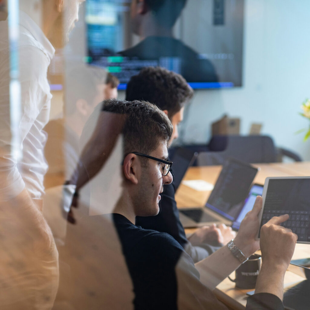 Coworkers working together at a conference table, with one person using a tablet and others using laptops in a bright office.
