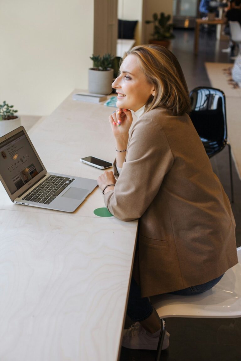 A woman with blonde hair wearing a tan blazer sits at a light wood desk with a laptop and smartphone, looking away while resting her chin on hand