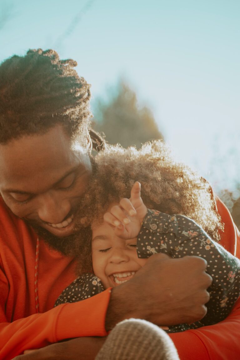 A smiling father hugs and playfully cuddles his laughing young child outdoors in warm sunlight.