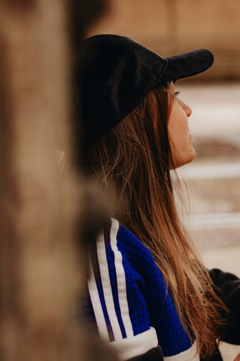 Side-profile portrait of a woman with long brown hair wearing a black baseball cap and a blue-and-white jacket, seated in warm indoor light.