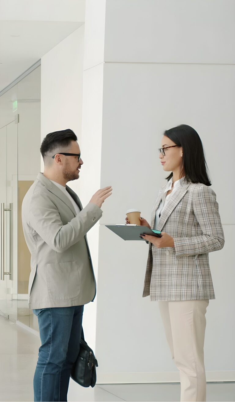 Two professionals wearing glasses converse in a bright office hallway; the man gestures while the woman holds a clipboard and takeaway coffee cup.