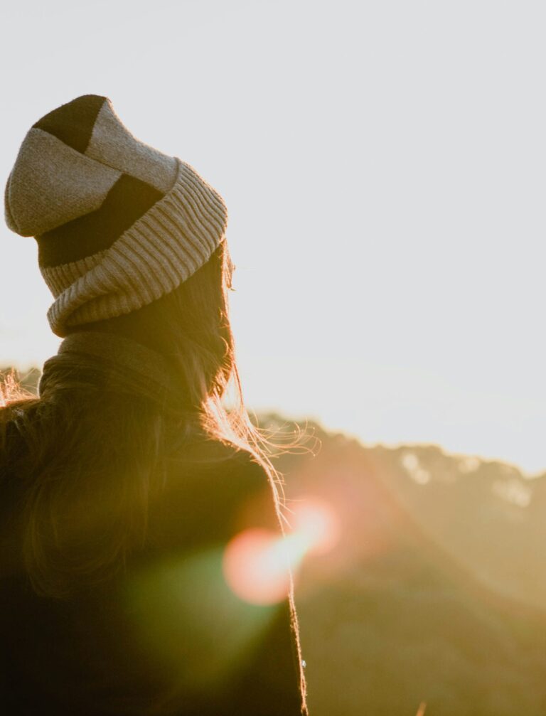 Person wearing a striped knit beanie and dark jacket, viewed from behind in warm sunset light.