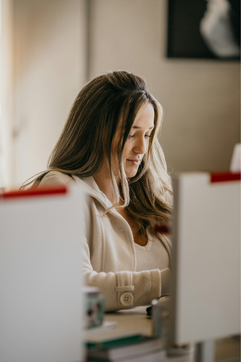Woman with long brown hair seated at a desk, looking down as she works between two computer monitors in a softly lit office.