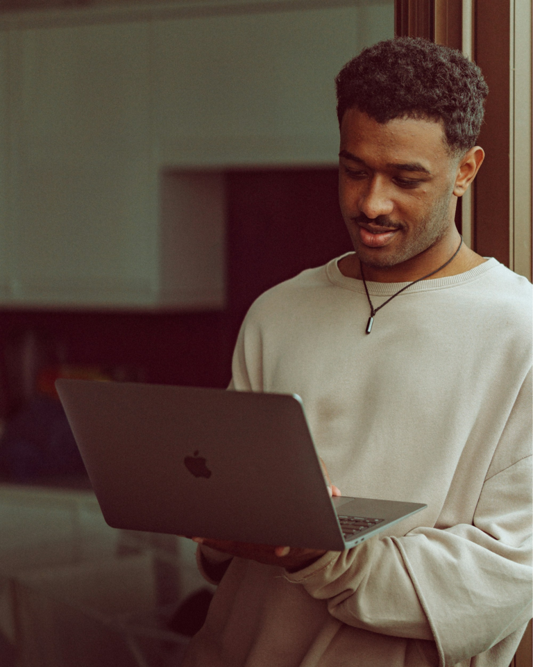 Man standing indoors and looking at an open laptop he holds in both hands.