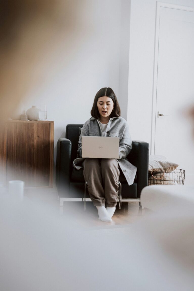 A woman sits in a chair using a laptop in a bright, minimalist room with neutral decor and a wooden cabinet in the background.